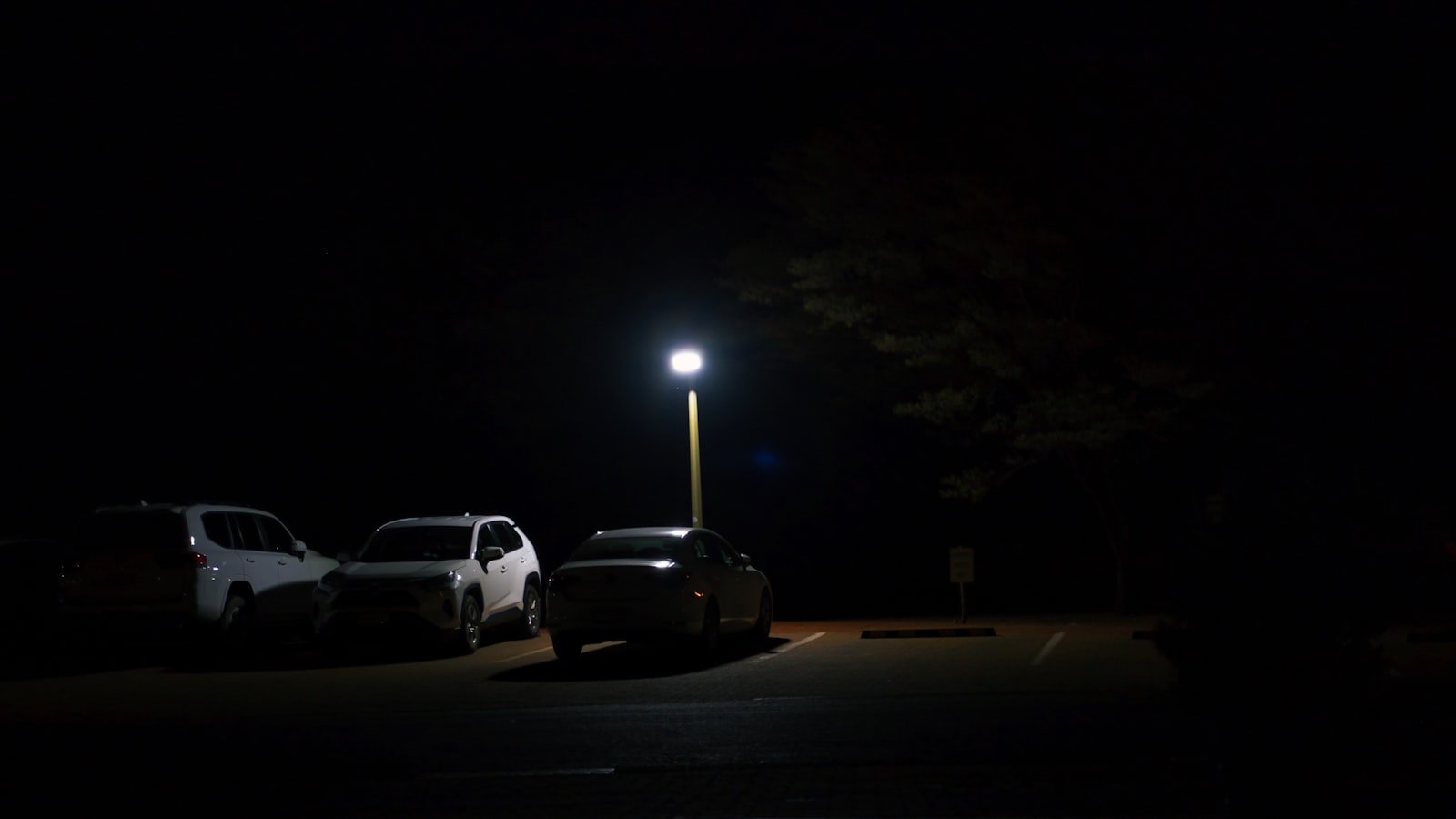 An empty parking lot at night lit by a single streetlight, with a few cars parked in the shadows